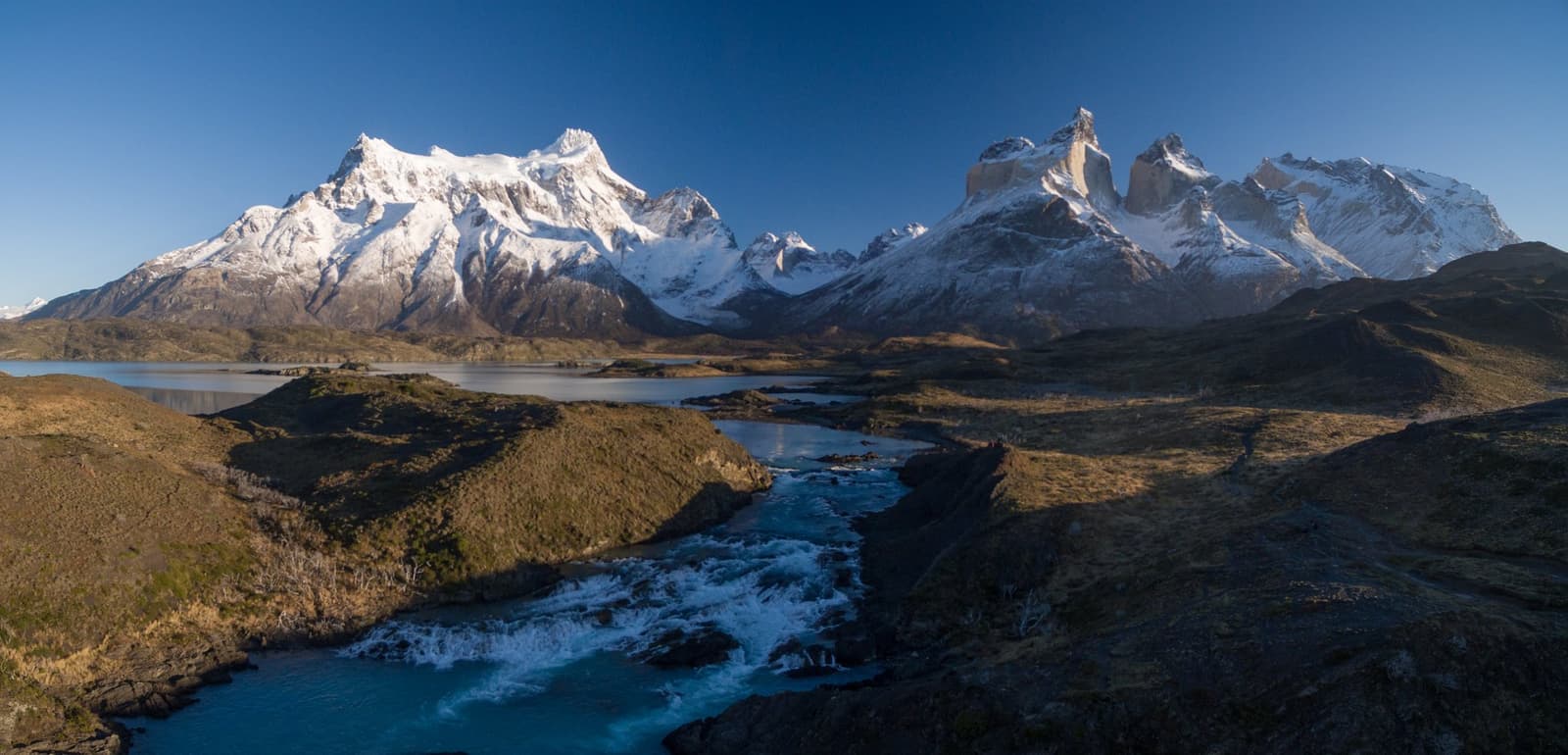 A river this colour needs no filter, just patience and good boots. Photo: Patagonia Camp Panoramic aerial view of a turquoise glacial river cascading through rocky terrain toward a calm lake, with the snow-capped peaks of the Paine Massif and the distinctive granite horns of Los Cuernos rising under a deep blue sky — the landscape surrounding Patagonia Camp