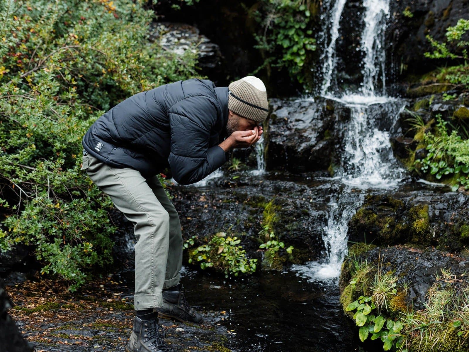 Cold water, no complaints. Some rituals are better without a bathroom. A hiker in a puffer jacket and beanie splashing cold waterfall water on his face beside a moss-covered cascade during a patagonia camp excursion