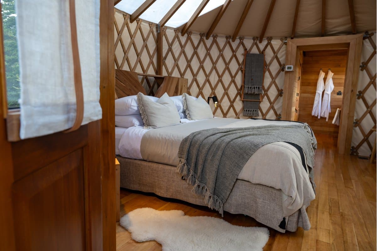 The bed is ready, the bathrobes are waiting, and the only decision tonight is which pillow to claim first. Photo: Patagonia Camp Interior of a circular patagonia camp yurt showing a king-size bed dressed in white linen and a grey fringed throw, a fluffy sheepskin rug on hardwood floors, latticed canvas walls, a skylight dome, and white bathrobes hanging near the ensuite door