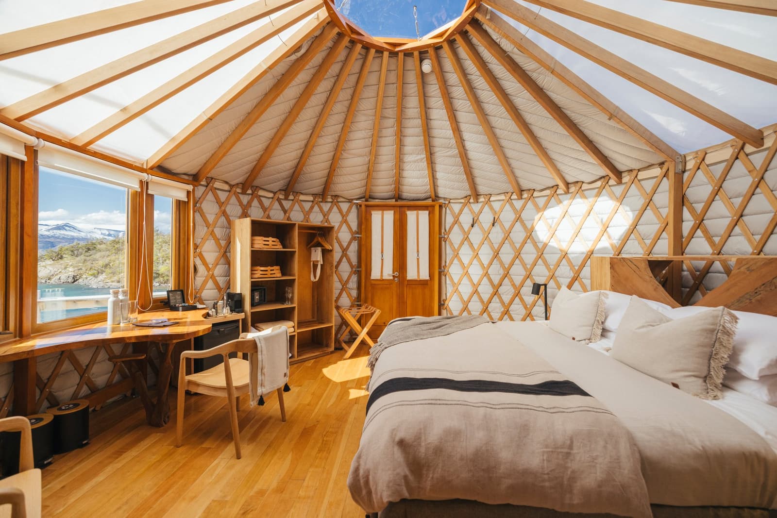 Round walls, wood beams overhead, and a snow-capped mountain just out the window, not a bad place to sleep. Photo: Patagonia Camp Interior of a patagonia camp yurt with a king-size bed dressed in neutral linen, warm wood lattice walls, a skylight crown, and a window framing a snow-capped mountain and turquoise lake beyond