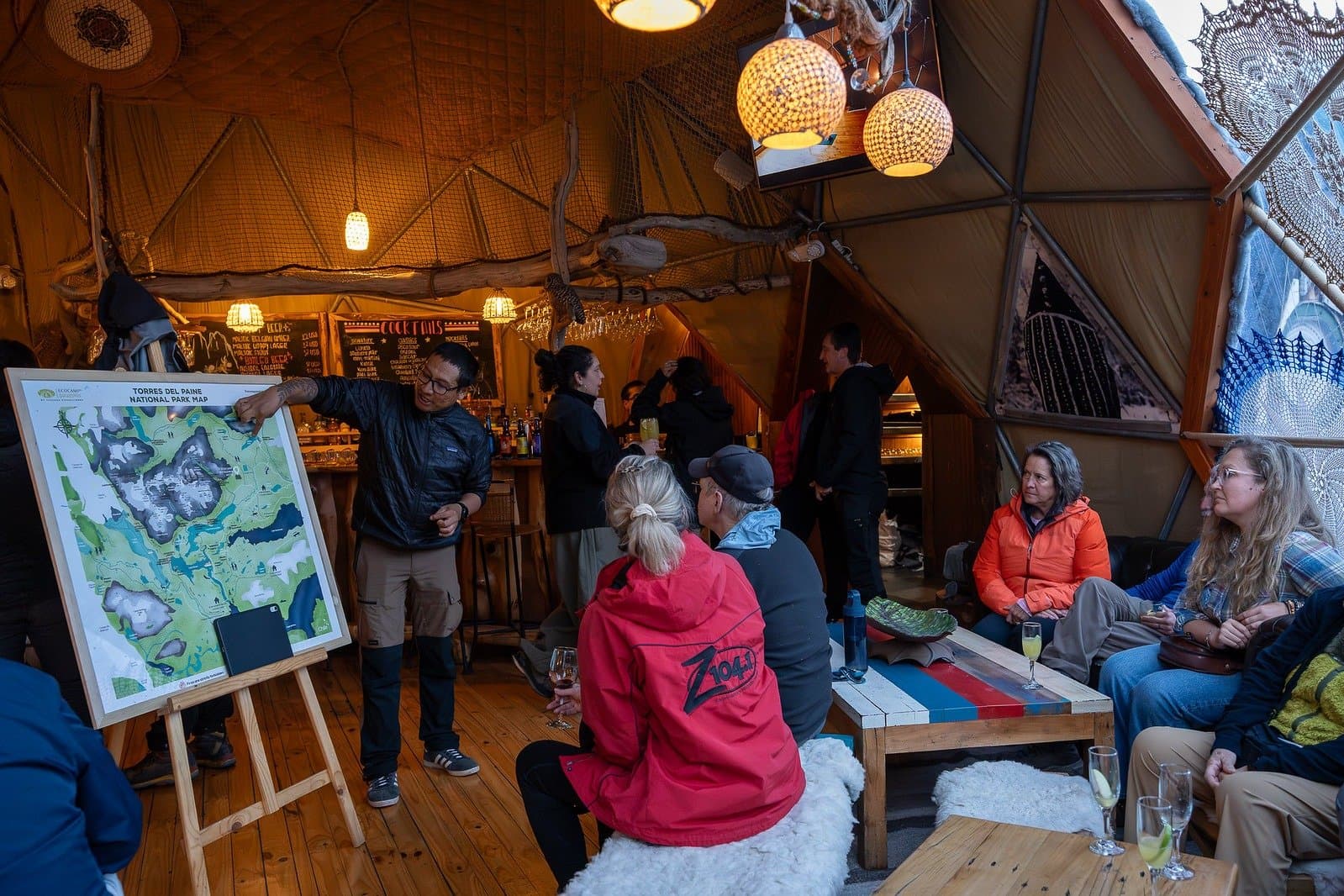 Before the pumas, the plan. Guides walk you through every corner of the park over a welcome drink. A guide points to a Torres del Paine National Park map on an easel during a puma trekking Patagonia welcome briefing, as guests relax with drinks inside a warmly lit geodesic dome tent at EcoCamp Patagonia