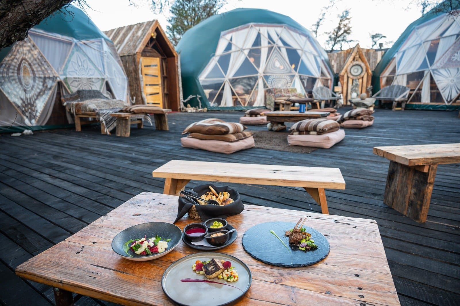 Camp food that makes you forget you spent the day scrambling across a windswept steppe. Multiple gourmet dishes plated on slate and ceramic served on a rustic wooden table on the outdoor deck of EcoCamp Patagonia, with decorated geodesic domes, floor cushions, and wooden benches visible in the background during puma trekking Patagonia expeditions