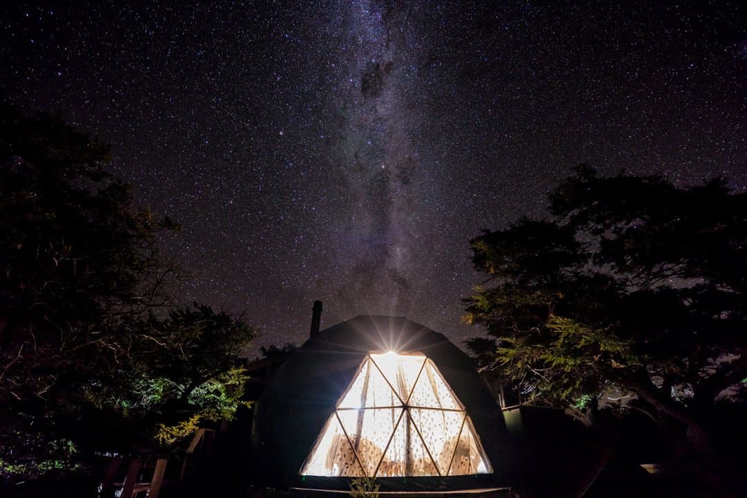 The dome is warm, the galaxy is free, and tomorrow the pumas are waiting. A glowing geodesic dome at EcoCamp Patagonia lit from within at night, surrounded by silhouetted trees, with the Milky Way stretching across the dark sky above