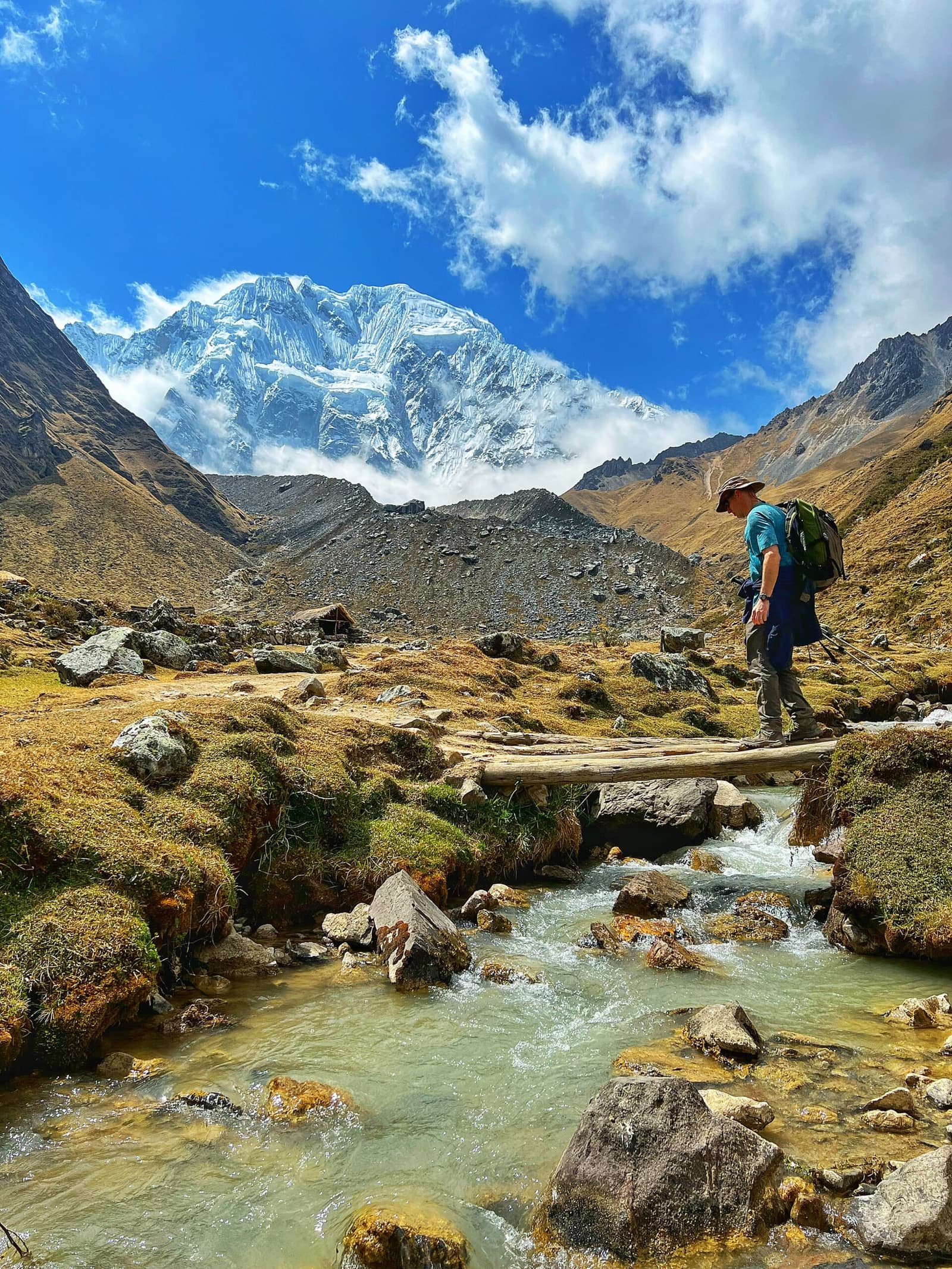 Um tronco, um riacho, um glaciar pela frente. Bom dia, Dia 2. Um trekker com mochila atravessa uma ponte rústica de troncos sobre um riacho de montanha cristalino no trek Salkantay de 5 dias, com um pico nevado imenso surgindo atrás do vale andino