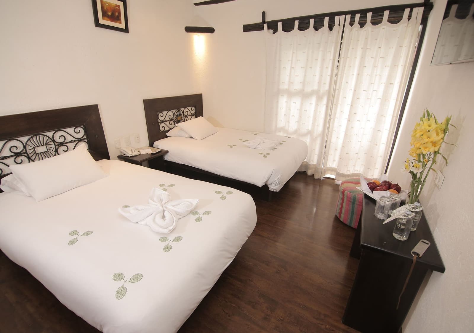 Lençóis limpos, frutas na mesinha e uma cama de verdade para descansar antes de Machu Picchu. Quarto duplo de hotel com cabeceiras de ferro ornamentadas, roupa de cama branca bordada com folhas, toalha dobrada em borboleta, vaso de flores amarelas e fruteira na mesa de cabeceira, no hotel em Aguas Calientes durante o trek Salkantay de 5 dias