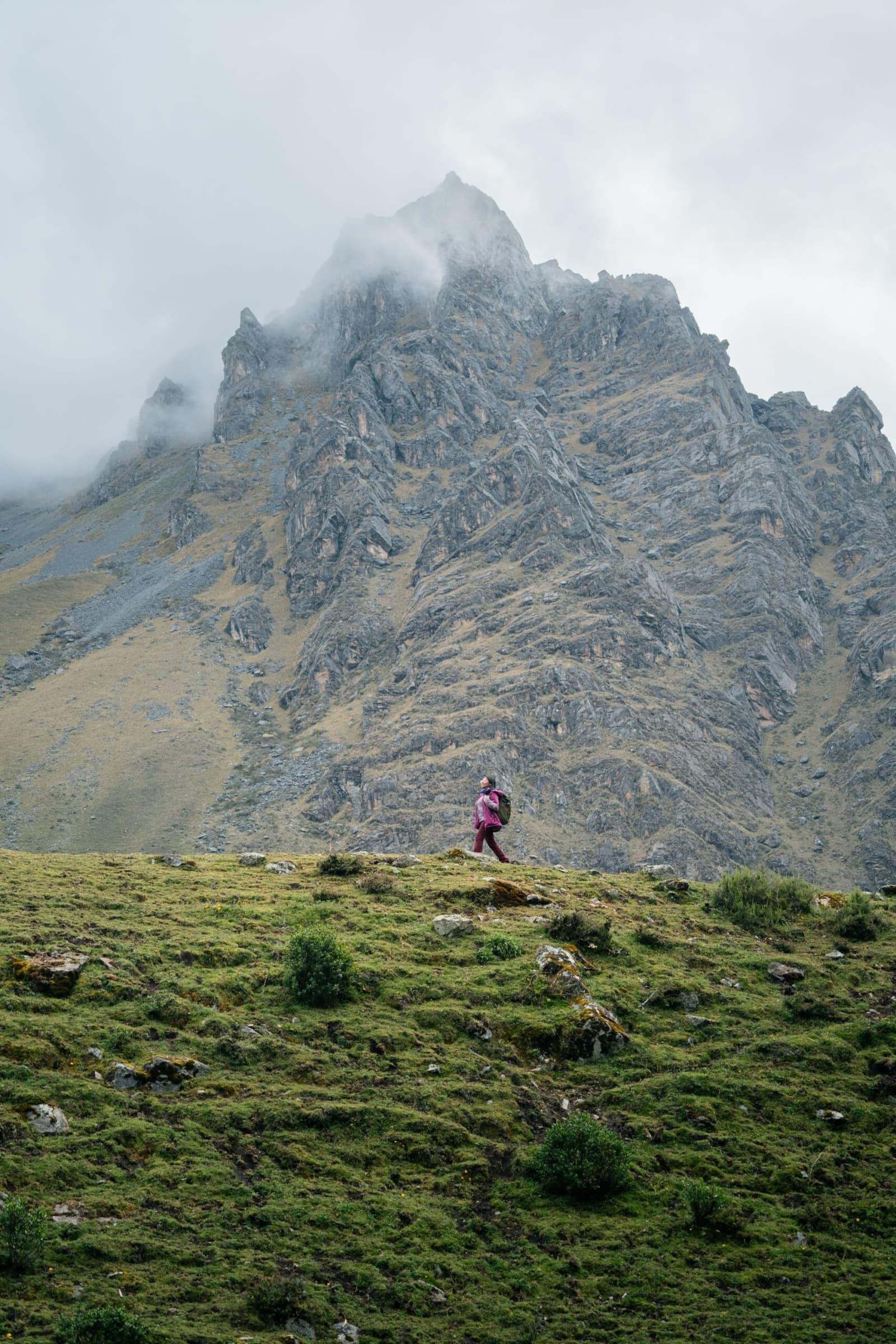 Uma pessoa, uma montanha, e a certeza de que você é menor do que pensa. Uma caminhante de jaqueta rosa e mochila atravessa uma campina alpina verde durante o trek Salkantay de 5 dias, com um pico rochoso recortado envolto em nuvens baixas ao fundo