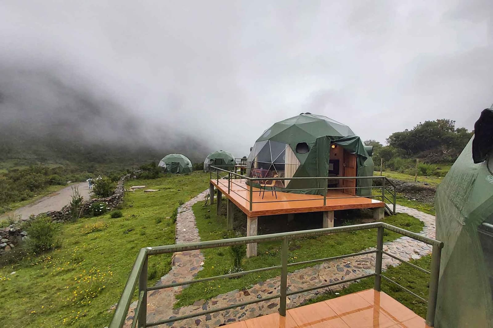 As nuvens chegam sem avisar, e ficam. Domos geodésicos verdes em decks de madeira elevados num acampamento de altitude na trilha Salkantay de 5 dias, com nuvens baixas cobrindo as colinas ao redor