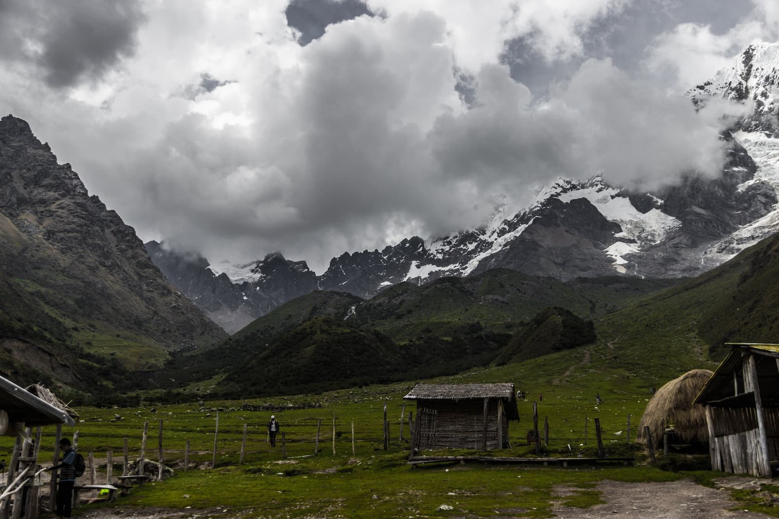 Tempestade chegando, geleiras firmes no lugar. A caminhada continua. Dois caminhantes atravessam um vale verde de alta altitude na trilha Salkantay para Machu Picchu, com abrigos rústicos de madeira em primeiro plano e picos nevados emergindo entre nuvens de tempestade