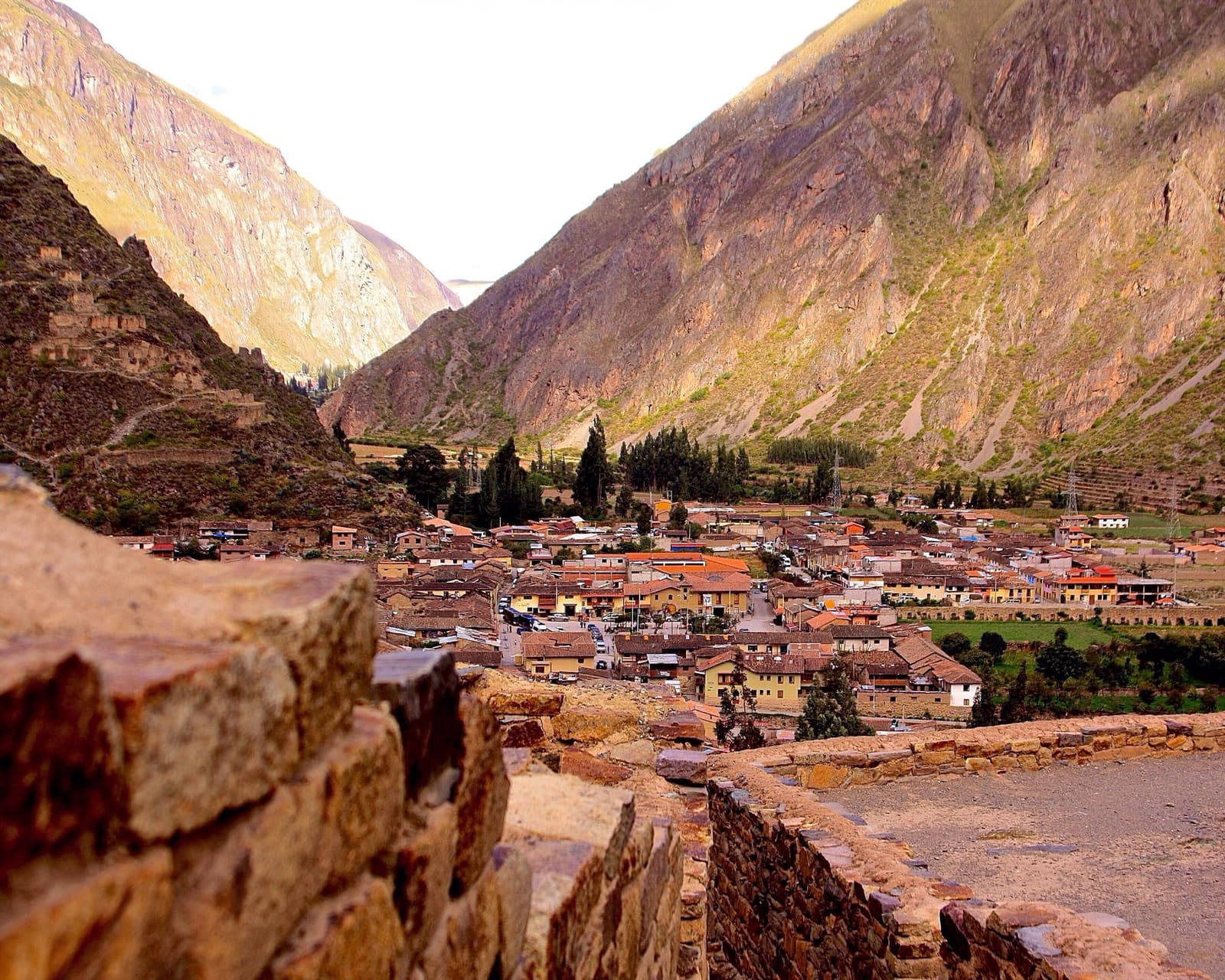 As mesmas pedras, o mesmo vale, quinhentos anos sem mudar a paisagem. Vista sobre a cidade de Ollantaytambo com telhados de terracota no Vale Sagrado, emoldurada por muros de pedra antiga em primeiro plano e picos íngremes dos Andes em ambos os lados