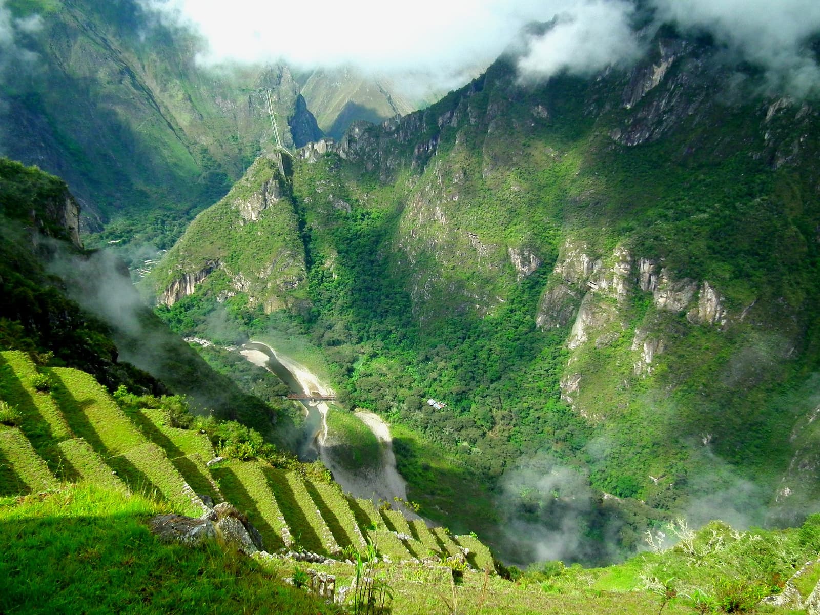 A névoa engole o cânion lá embaixo; os terraços ficam de pé, indiferentes. Vista aérea de antigas terraços agrícolas incas na trilha Salkantay até Machu Picchu, com névoa subindo do cânion do rio Urubamba e montanhas cobertas de floresta ao fundo