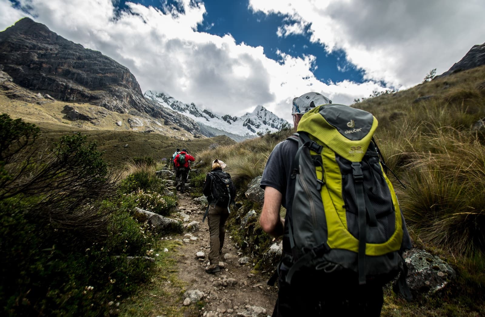One foot in front of the other, and suddenly the Andes open up. Three hikers with trekking packs walking single file on a rocky trail through high-altitude puna grassland on the santa cruz trek, with a snow-capped peak and dramatic clouds visible in the valley ahead