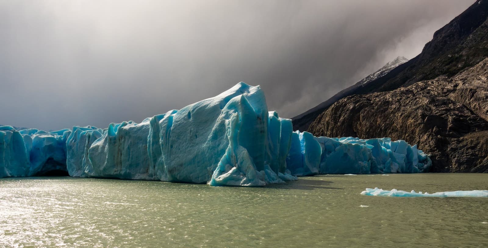 That blue is ancient compressed ice, and it sounds different up close. Towering blue ice walls of Grey Glacier meet a grey-green glacial lake on the self-guided W Trek, with dark storm clouds and a rocky Patagonian mountainside behind
