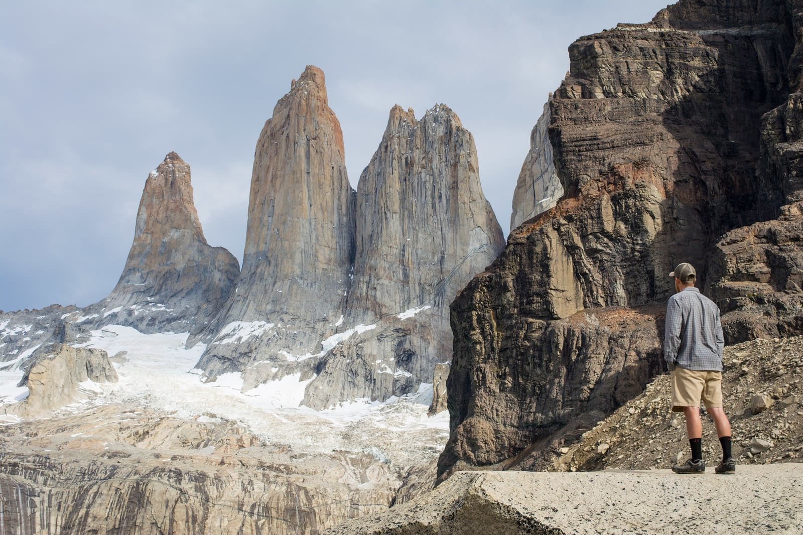 One boulder field, three towers, zero regrets. A hiker in a cap and shorts stands at the base of the towers on the short W trek, looking up at the three granite spires rising above a snow-covered moraine under a cloudy Patagonian sky