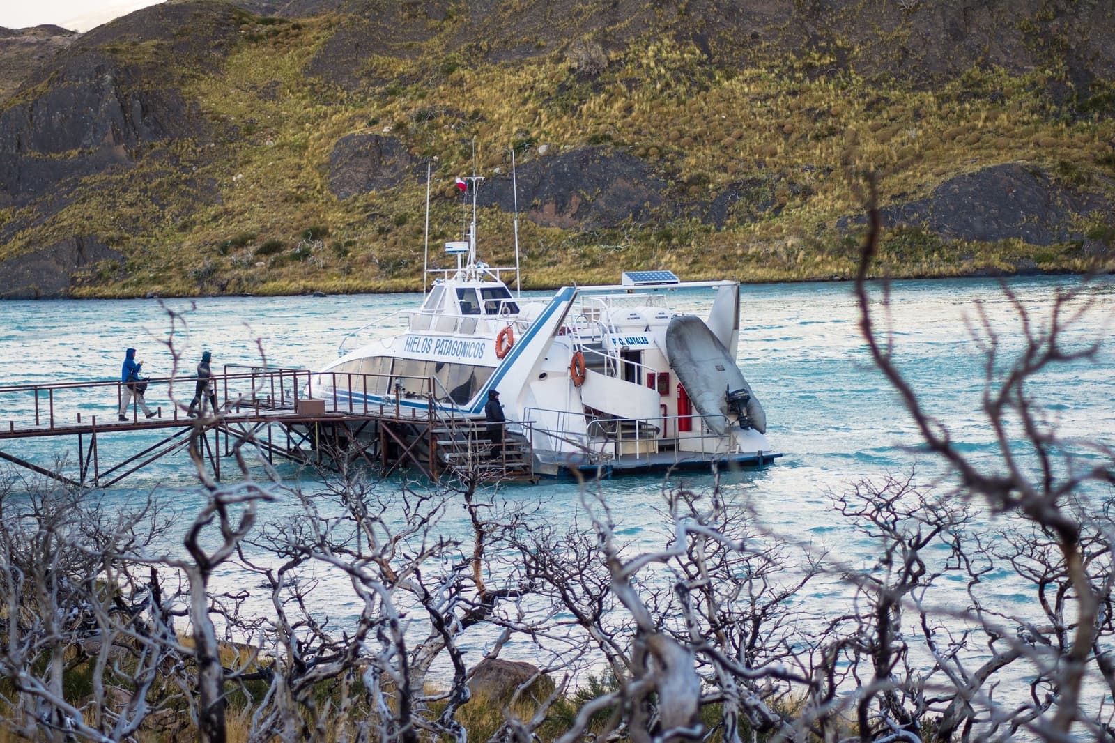 Bare branches, turquoise water, and a catamaran that means the trek has officially begun. The Hielos Patagónicos catamaran docked at a metal pier on turquoise Lake Pehoé, framed by bare lenga beech branches in the foreground, as passengers board for the short W trek crossing to Paine Grande