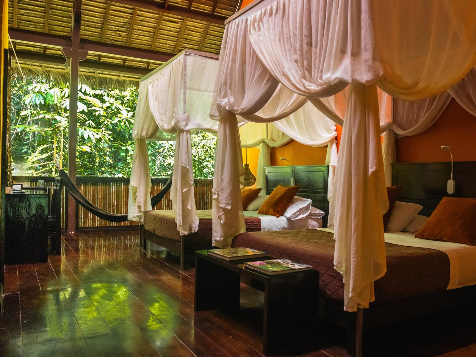 A hammock on the porch, the forest right there, no wall between you and it. Interior of a Tambopata Research Center Peru lodge room featuring two four-poster beds with white canopy drapes, warm terracotta walls, dark hardwood floors, a hammock on the open veranda, and lush rainforest greenery visible beyond the open-front bamboo railings