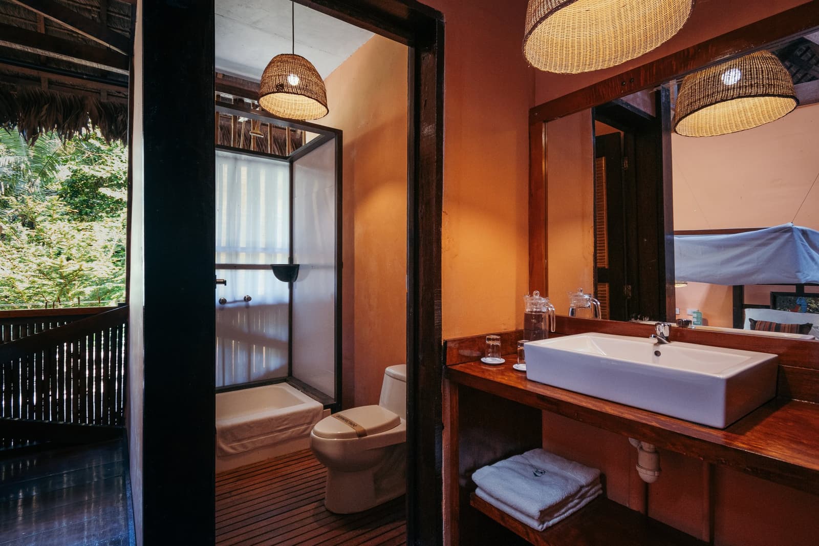 Warm terracotta walls, proper towels, and the forest right outside the window. Photo: GABRIEL_S Private en-suite bathroom at the Tambopata Research Center Peru lodge, featuring a vessel sink on a dark wood vanity, wicker pendant lights, and a glass-enclosed shower, with lush rainforest canopy visible through the open-air window.