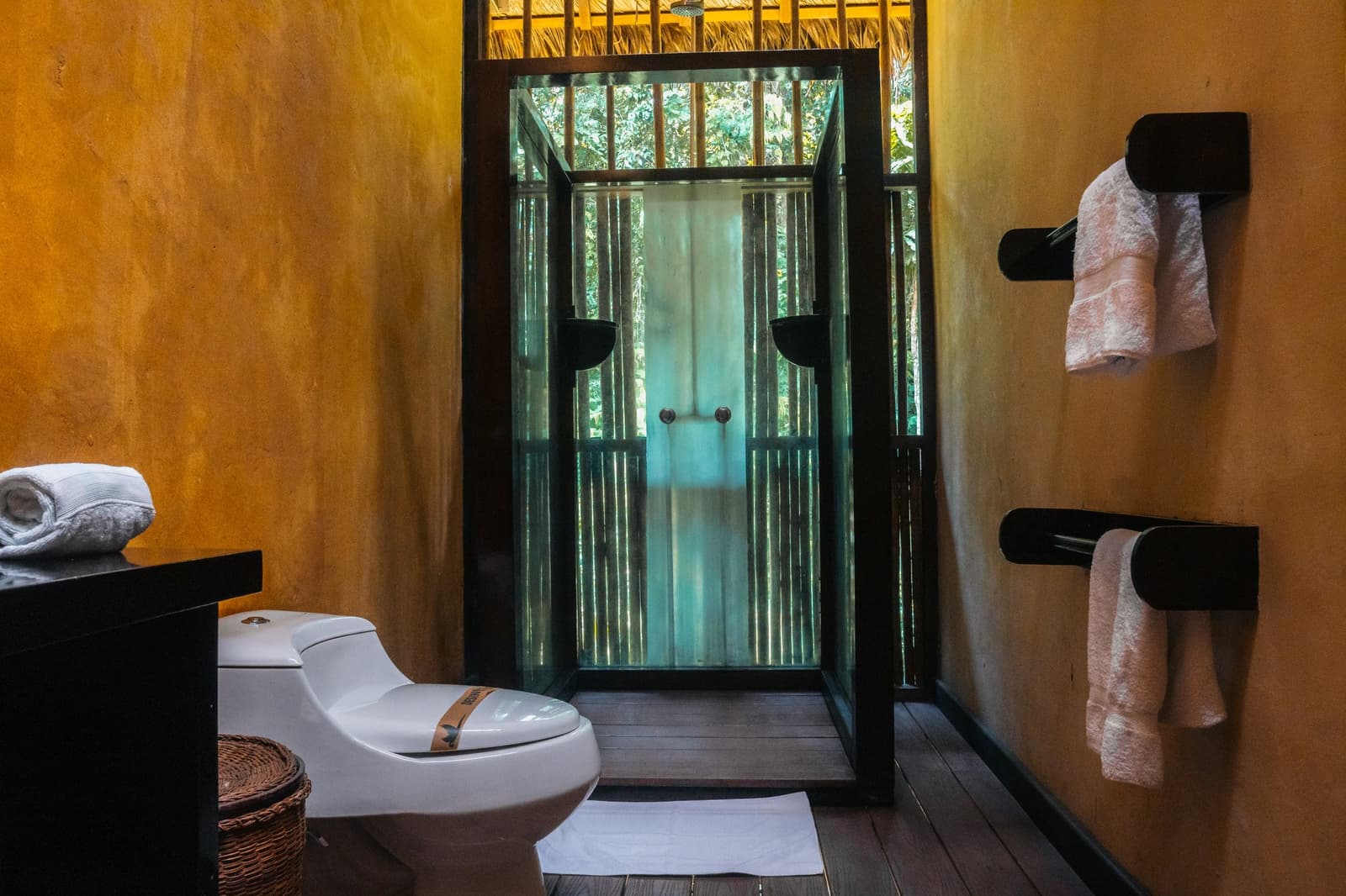 Hot shower, forest canopy overhead. Roughing it was never part of the deal. Private bathroom at the Tambopata Research Center Peru lodge, featuring ochre-plastered walls, a glass-enclosed shower with forest views through the open roof, black timber accents, folded towels on wall-mounted racks, and a wicker bin on a dark hardwood floor