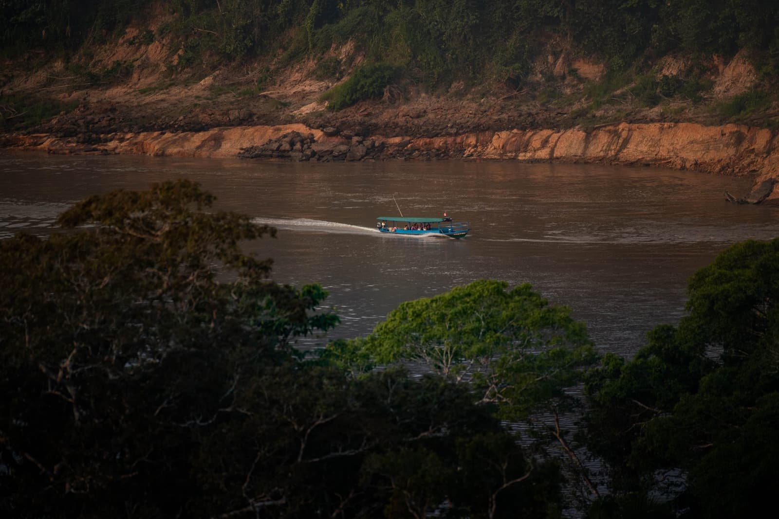 Three hours upriver, the last houses disappear and the forest takes over completely. A blue passenger boat cruises up a wide Amazonian river toward the Tambopata Research Center Peru, framed by forest canopy in the foreground and a red clay riverbank rising behind it in warm late-afternoon light.