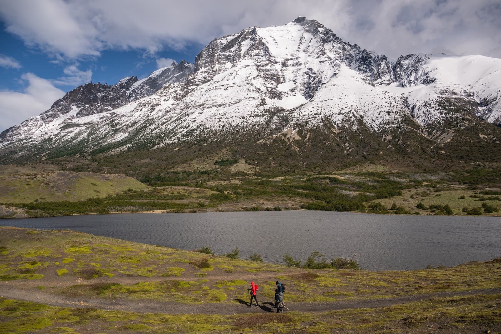 Small figures, enormous mountain. The scale hits you mid-stride. Two hikers walk a trail beside a calm alpine lake on a torres del paine 4 day all inclusive tour, with a snow-covered granite peak rising steeply behind them under a partly cloudy sky