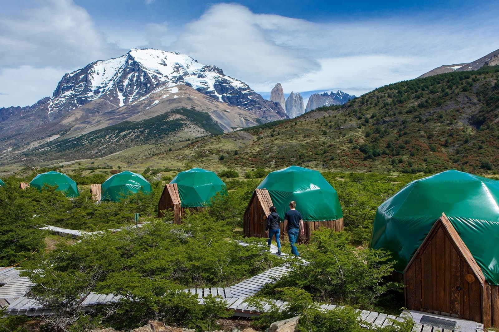 Your camp, your domes, and those towers keeping watch from the ridge. Two guests walk a wooden boardwalk past a row of green geodesic domes at EcoCamp Patagonia, with the Torres del Paine 4 day all inclusive tour base framed by snow-capped peaks and the iconic granite towers in the background