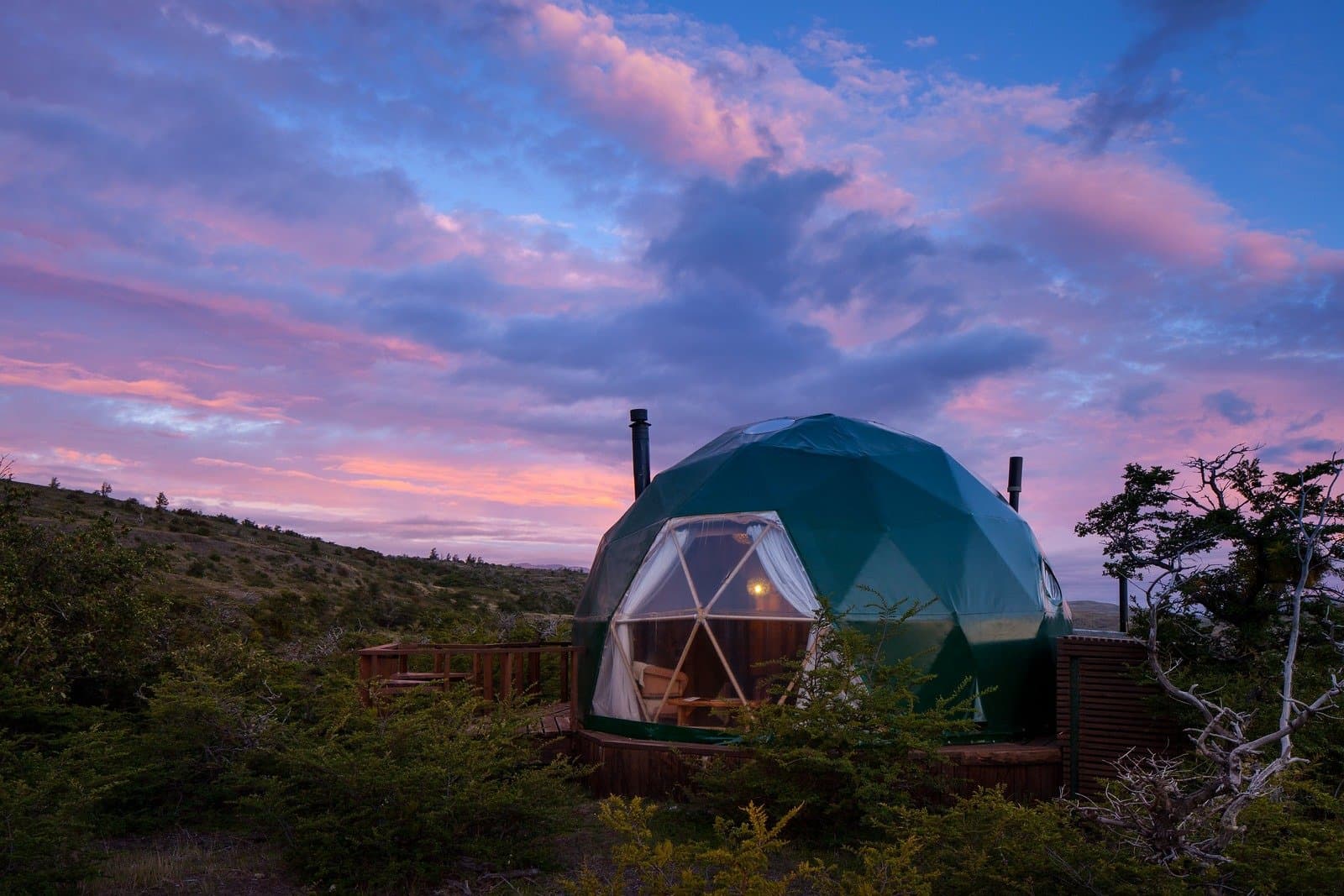 Pink sky, warm light inside, and nothing but Patagonian steppe in every direction. A green geodesic glamping dome at EcoCamp Patagonia glows warmly from within at dusk, set among low scrub and windswept trees beneath a pink and blue twilight sky on this torres del paine 4 day all inclusive tour