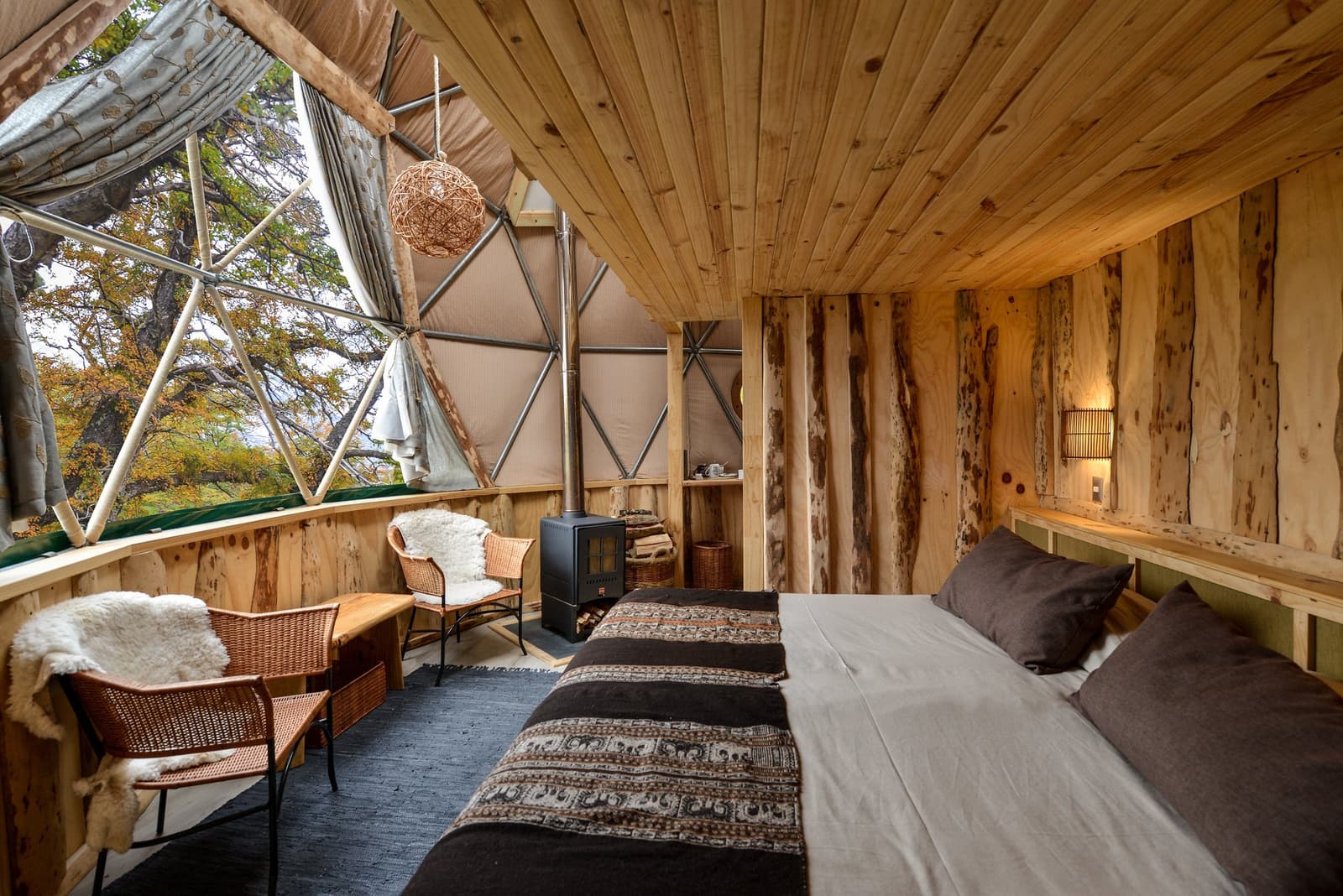 Lenha no fogo, pele de ovelha na cadeira, e a Patagônia lá fora esperando você. Photo: Suite Dome Loft Interior de um domo geodésico na Torres del Paine W Trek, com fogão a lenha, cadeiras de vime cobertas com pele de ovelha, paredes de madeira rústica e uma grande janela triangular com vista para árvores no outono