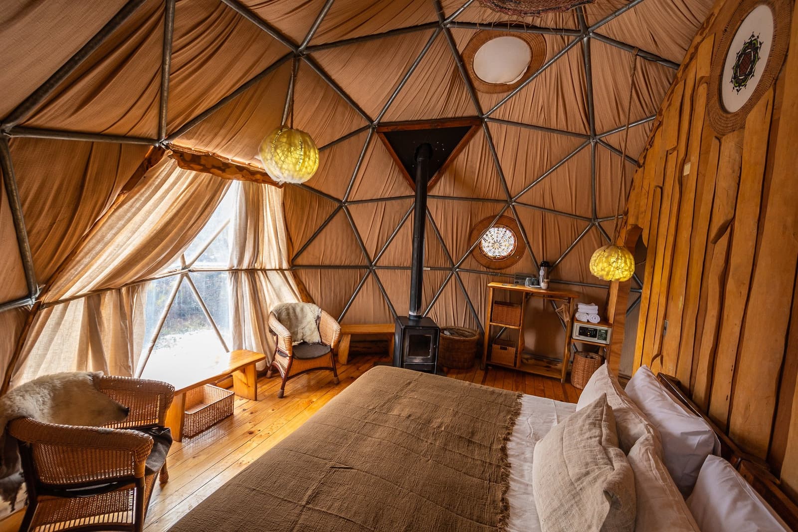 Fogão a lenha, pele de ovelha e luz âmbar, acampar nunca foi tão relativo. Photo: Suite Dome Interior de uma cúpula geodésica no trekking W de Torres del Paine, com cama de casal, fogão a lenha preto, luminárias globo âmbar, cadeiras de vime com peles de ovelha e assoalho de madeira aquecido
