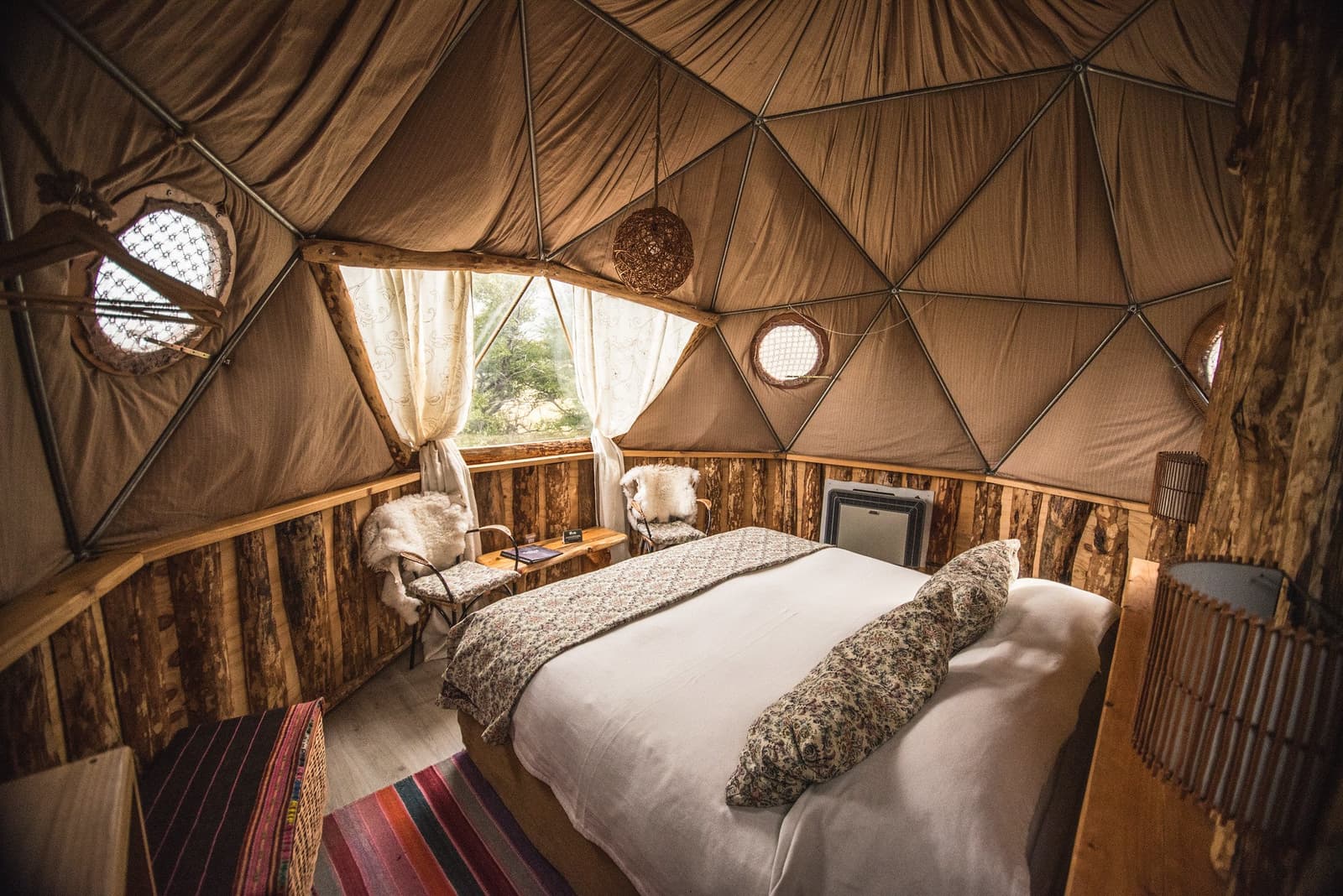 Depois de uma noite assim, sair para a trilha exige uma força de vontade extra. Photo: Superior Dome Interior de um domo geodésico no Trek W Torres del Paine, com cama de casal em roupa de cama floral, cadeiras com pele de carneiro, paredes de troncos rústicos, janelas redondas tipo vigia e luminária pendente trançada