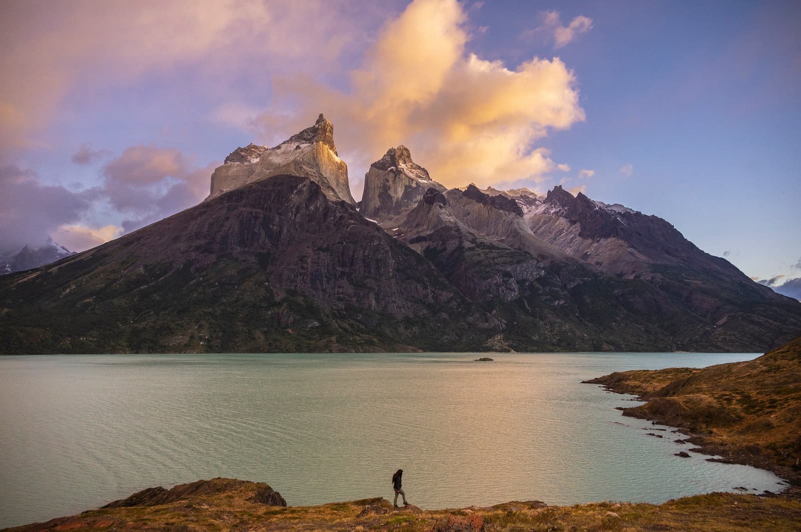 Small figure, enormous mountain. Scale has a way of putting things in perspective. A lone hiker walks along the glacial turquoise shore of a Patagonian lake on the Torres del Paine W Trek, with the dramatic granite horns of the Paine Massif and golden clouds filling the sky above