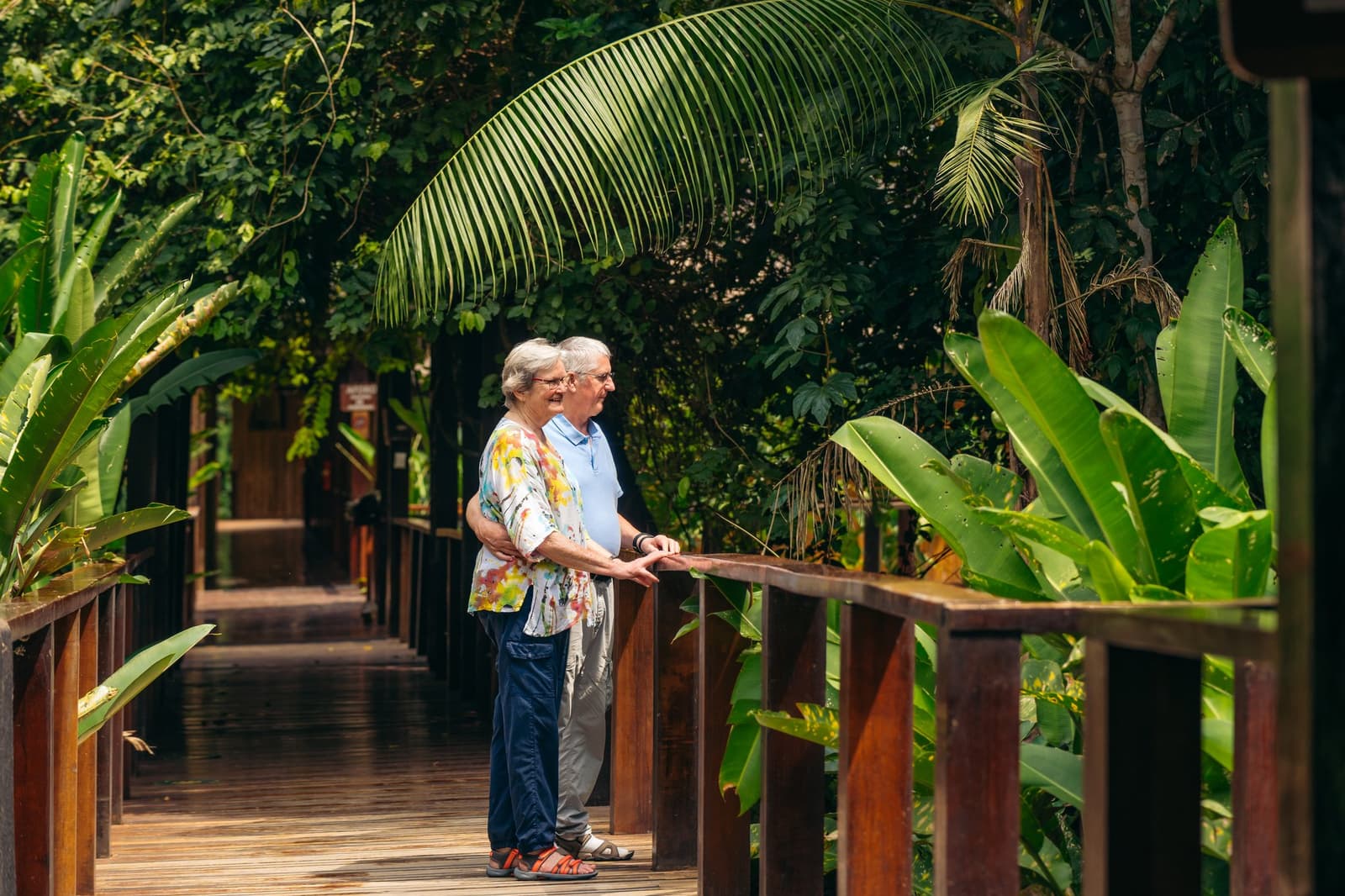 Two people, one railing, and a whole lot of jungle to talk about. An older couple stands arm in arm on a wooden boardwalk at Posada Amazonas, leaning against the railing and looking out into dense tropical foliage on this amazon tour from Puerto Maldonado