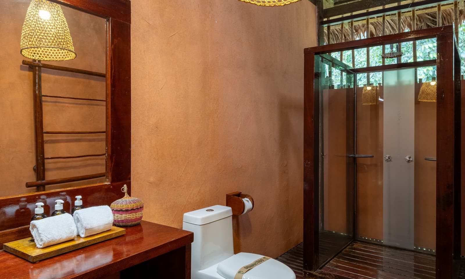 Hot shower, fresh towels, and the Amazon just outside the glass. Private en-suite bathroom at Posada Amazonas lodge, featuring a clay-toned plastered wall, dark hardwood vanity with rolled white towels and liquid soap dispensers, a woven basket accessory, bamboo-framed mirror, wicker pendant lamp, and a glass-panel shower cabin with a thatched roof and jungle foliage visible beyond the open upper wall.