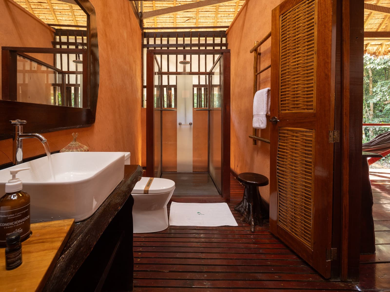 Mud walls, rain shower, and a hammock waiting just outside the door. Private bathroom at Posada Amazonas lodge featuring a vessel sink on a dark wood vanity, glass-enclosed rain shower, slatted hardwood floors, terracotta walls, and a louvred door opening to a jungle view with hammock — part of this amazon tour from puerto maldonado