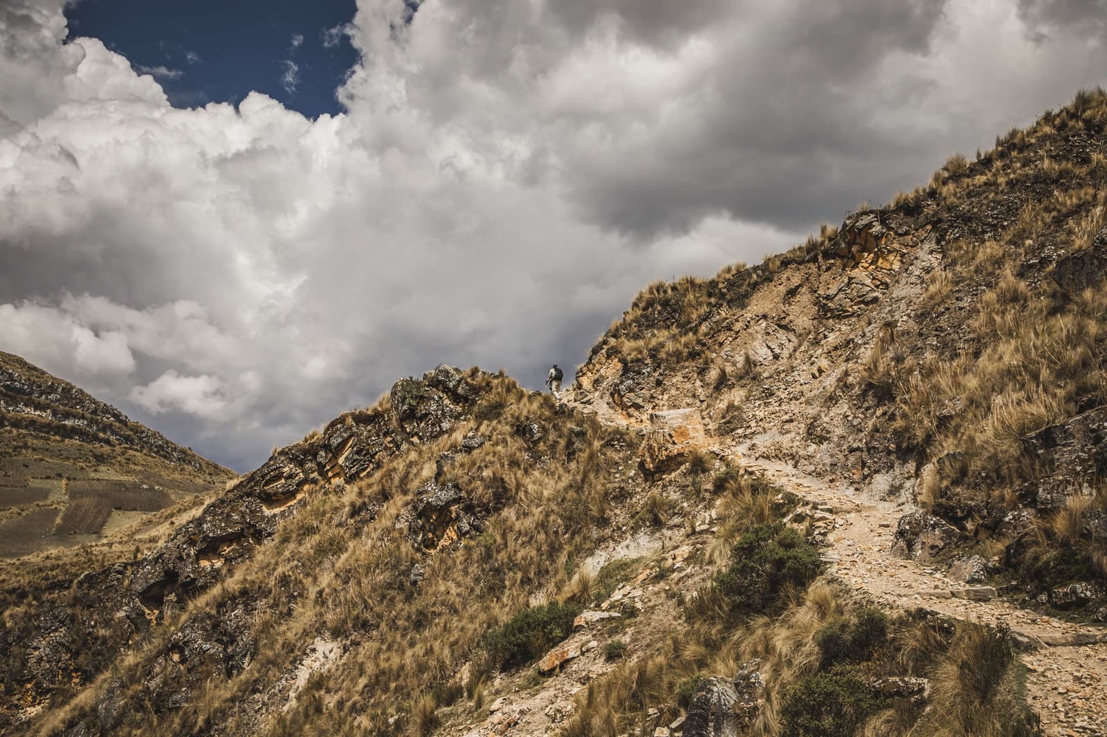 Lá em cima, é a trilha que manda no ritmo. Um caminhante solitário com mochila sobe uma crista rochosa coberta de gramíneas na Ancascocha Trek, diminuído por um céu dramático de cúmulos imponentes sobre a paisagem andina de alta altitude