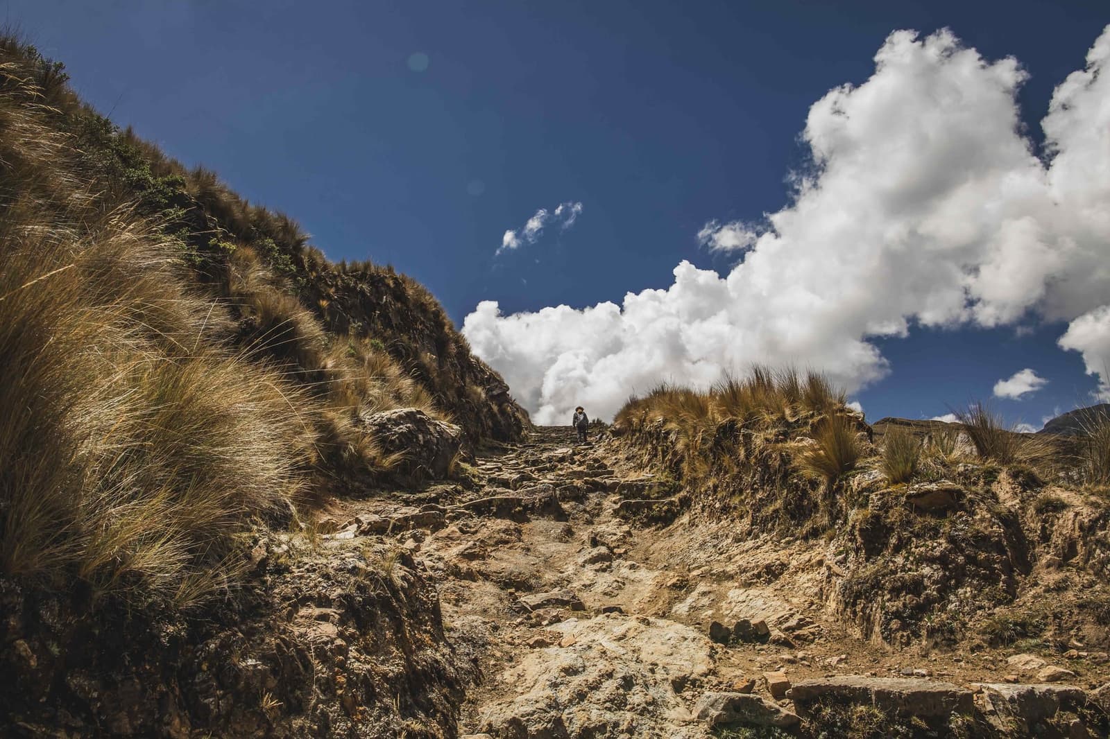 Uma figura pequena, uma subida longa. A altitude cobra o pedágio. Um caminhante solitário sobe um caminho rochoso e íngreme ladeado por capim ichu dourado na trilha Ancascocha, com grandes nuvens brancas no céu azul acima