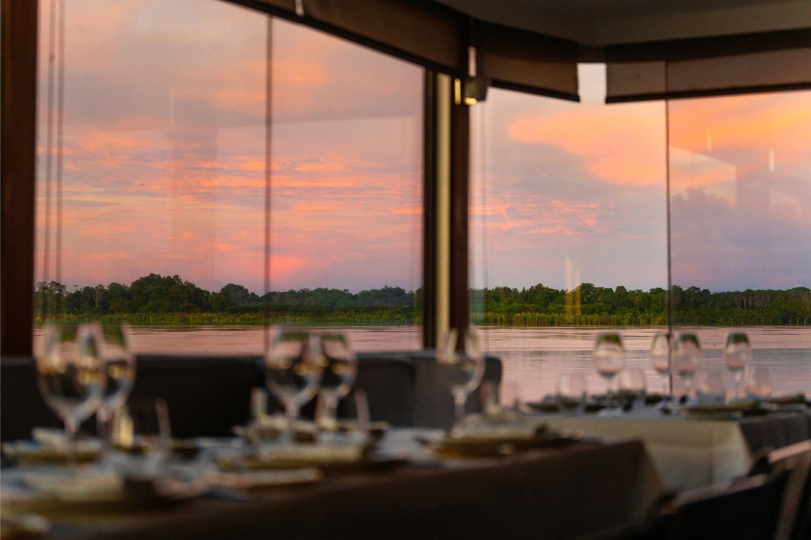 Dinner reservations: the river, the sky, and whatever the chef decides. Photo: Daniel Silvia Yoshisato Elegant dining room of the Aria Amazon cruise ship with wine glasses set on linen-draped tables and floor-to-ceiling panoramic windows overlooking a pink and orange sunset sky reflected on a calm river, with a dense treeline on the far bank