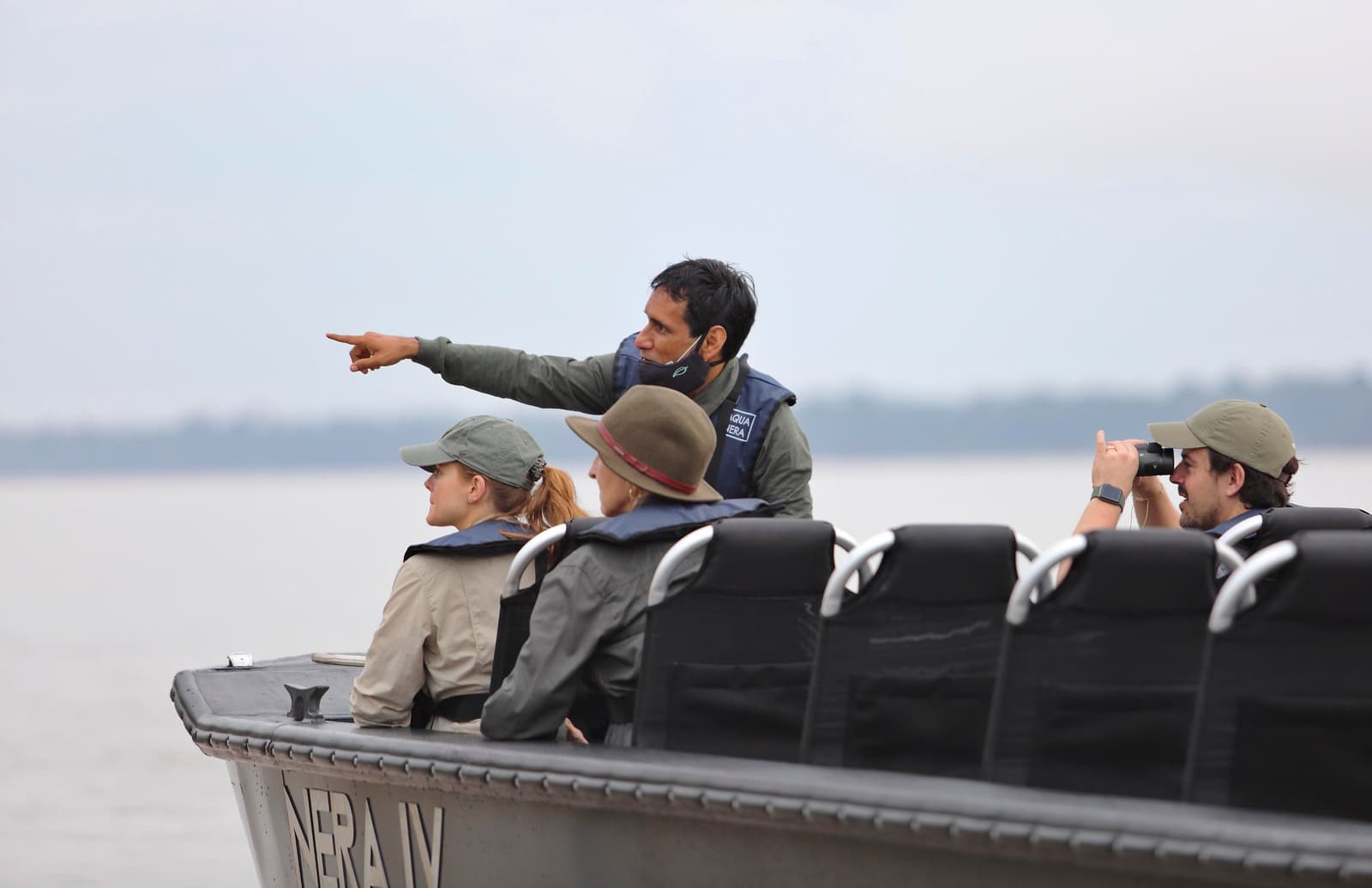 Four sets of eyes, one pointed finger, zero agreement on what it was. Photo: Stevie Mann A naturalist guide on an aria amazon cruise skiff points toward the riverbank while three guests in life vests scan the water, one using binoculars, on a wide hazy river