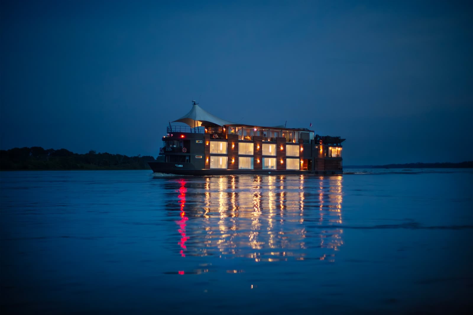 Out here, dusk is when the ship really comes alive. Photo: Daniel Silvia Yoshisato The Aria Amazon cruise ship glowing with warm interior lights reflects on the calm dark waters of the river at dusk, with a forested bank silhouetted against a deep blue sky