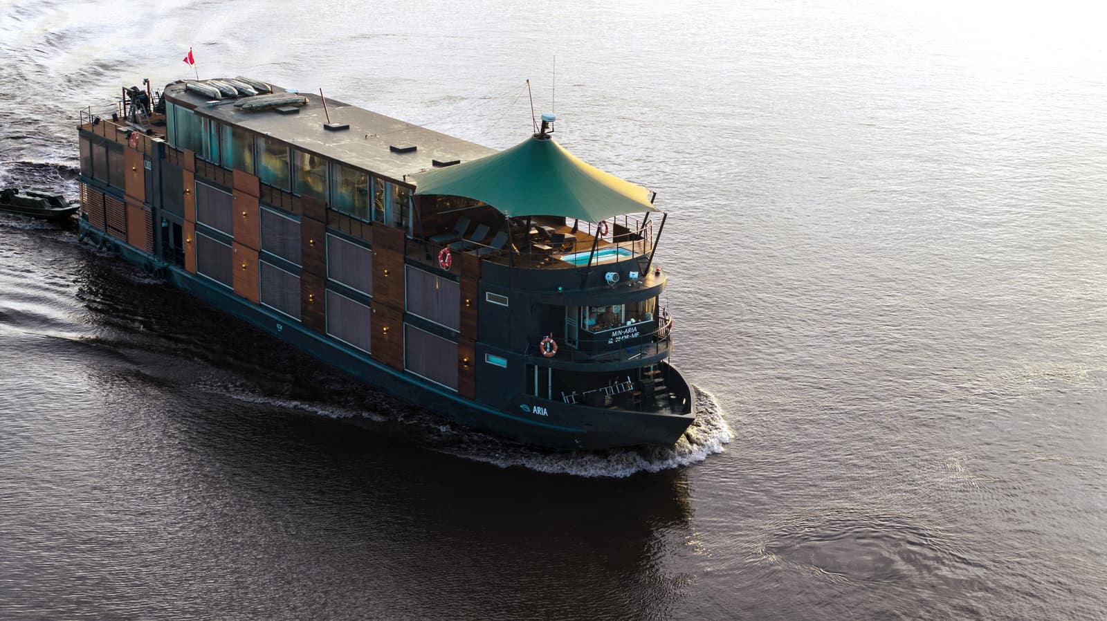 Thirty-two guests, one wide river, and a floating hotel that actually moves. Photo: Anthony K Do Aerial view of the Aria Amazon cruise ship navigating a wide river in Peru, with warm wood-paneled suites, a teal canopy over the open deck, and a small skiff alongside