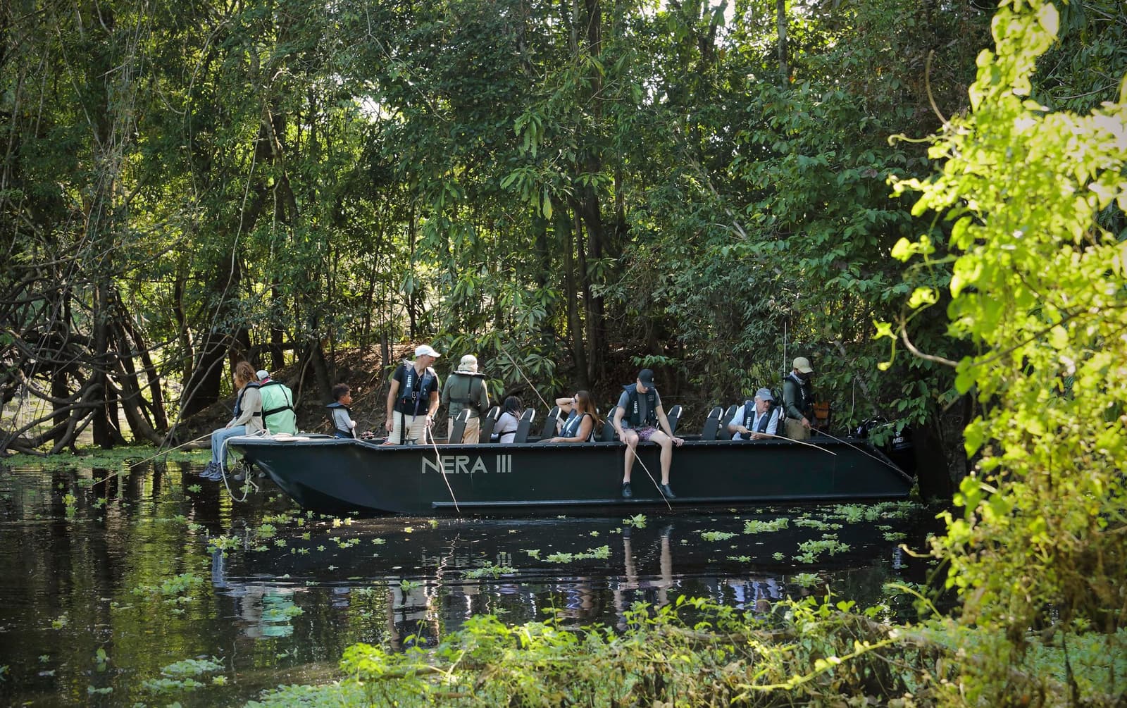 Fishing rods out, engines off, the forest doing all the talking. Photo: Stevie Mann A group of guests aboard the Nera III skiff on the aria amazon cruise, drifting through a narrow black-water channel lined with dense tropical forest, some standing with fishing rods and others seated at the bow