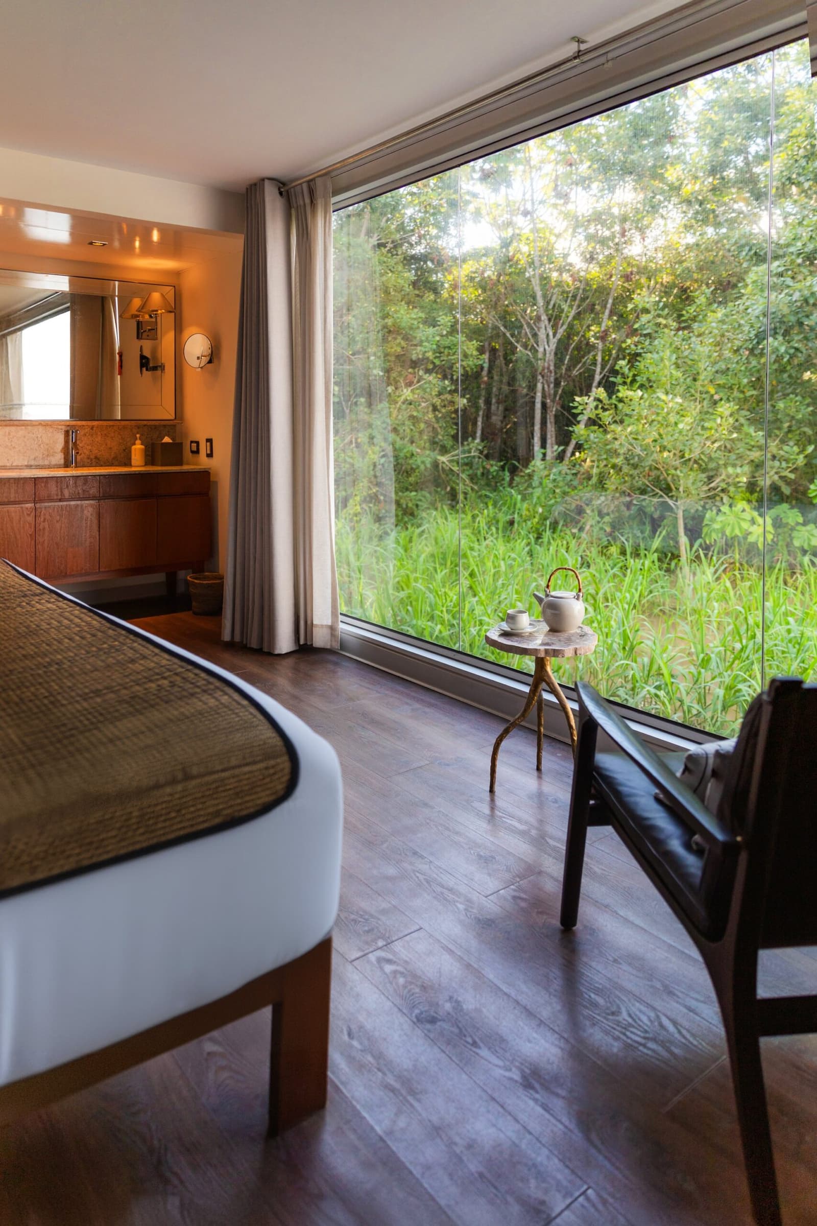 Tea for one, the jungle for company, not a bad morning ritual. Photo: Yvonne Kreuzmayr Aria Amazon cruise suite interior with a floor-to-ceiling glass wall overlooking dense green rainforest vegetation, a leather armchair, a small side table with a tea set, warm wood finishes, and a vanity area visible in the background