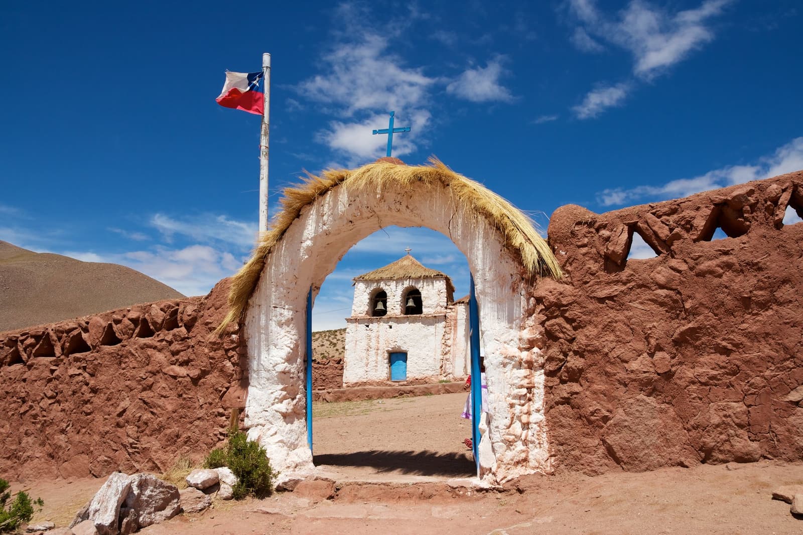La iglesia más sola del altiplano, y aun así la más visitada del camino. Una iglesia de adobe encalada con cruz azul y arco techado de paja ichu, rodeada de muros de piedra volcánica roja y una bandera chilena bajo un cielo azul intenso en el altiplano atacameño