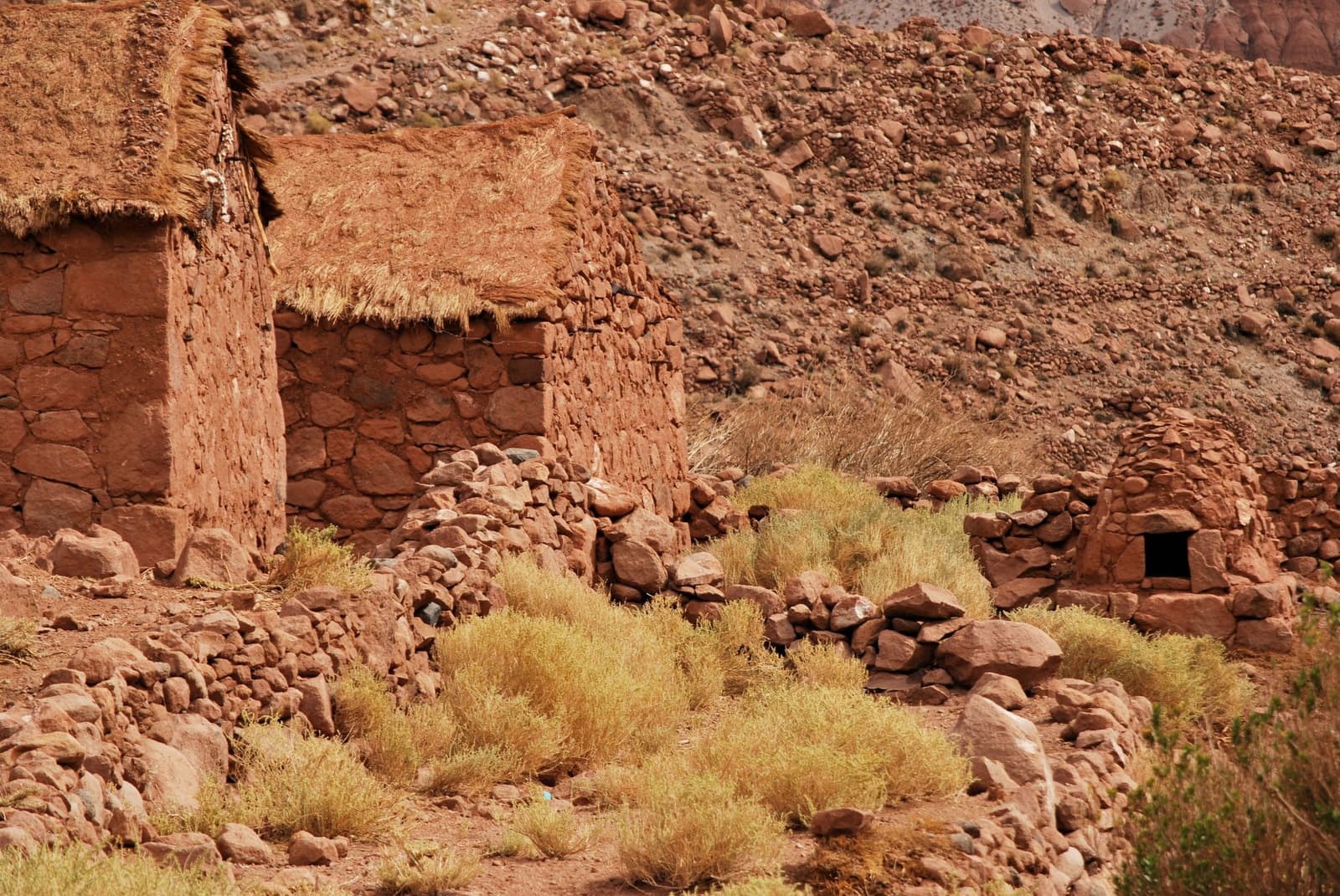 Hecha de la propia ladera, piedra a piedra, generación tras generación. Construcciones tradicionales de piedra roja y adobe con techos de paja en un paisaje árido de altura andina, rodeadas de arbustos secos y terreno rocoso