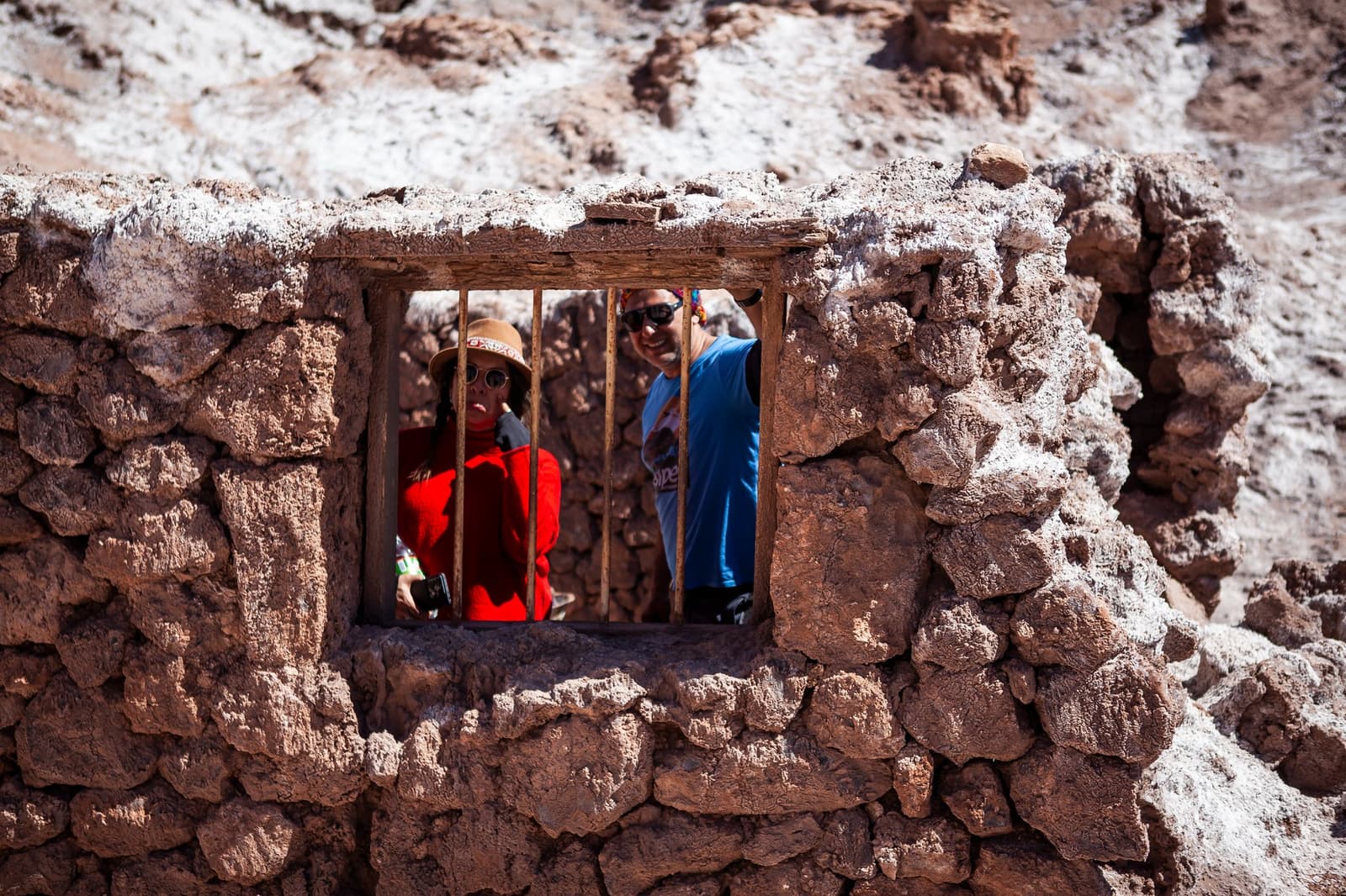 La mina lleva décadas cerrada, pero sigue siendo la mejor fotógrafa del valle. Photo: Mina Victoria Dos viajeros sonrientes asomados por una ventana enrejada en el muro de piedra erosionada y con costras de sal de las ruinas de la Mina Victoria en el Valle de la Luna