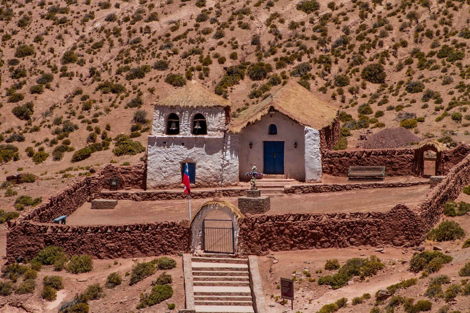 Mud, straw, stone, and a flag. Machuca has been here longer than the road that brought you. Photo: Iglesia Machuca Small whitewashed adobe church with thatched roofs and a bell tower, enclosed by a dry-stone wall and a Chilean flag, set against an arid hillside on an Atacama desert tour package stop at Machuca village