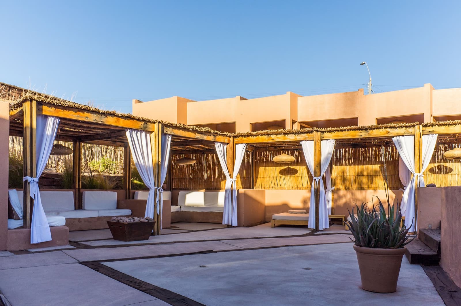 A spot made for doing nothing, and doing it well. Open-air lounge area of an Atacama desert tour package hotel, with white-curtained wooden pergolas, cushioned adobe benches, and a potted agave plant under a clear blue sky