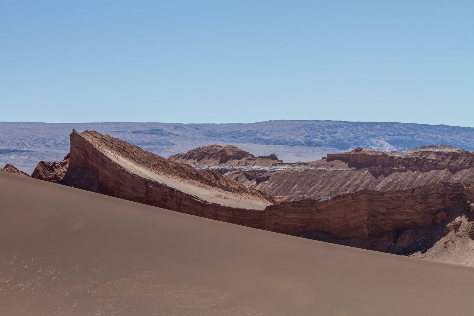 Sand and stone arguing over the same patch of desert, and both winning. Photo: Rodolfo GOUD A smooth sand dune foreground meets sharply eroded red and ochre rock ridges in Valle de la Luna, Atacama Desert, with a pale blue sky and distant plateau on this atacama desert tour package itinerary.