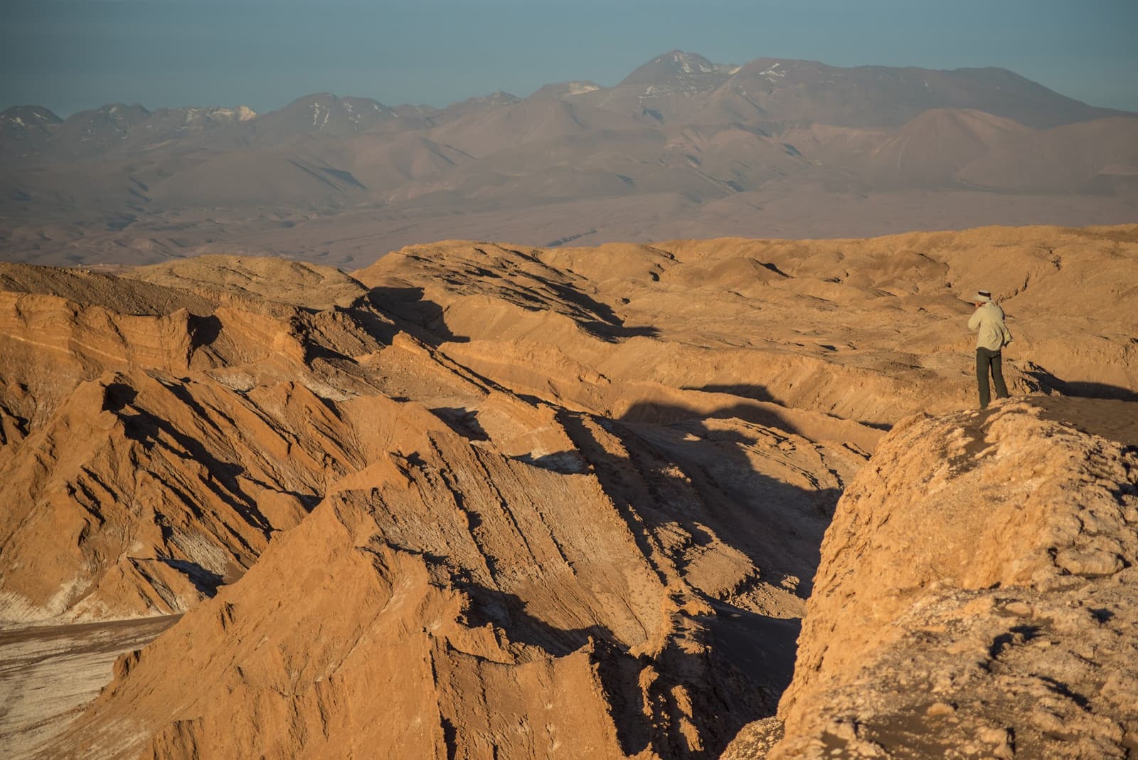 All that geology, and you still reach for your camera. A lone traveler stands on a rocky ridge during an atacama desert tour package, photographing the vast eroded salt mountain formations stretching toward snow-capped Andean peaks in the distance