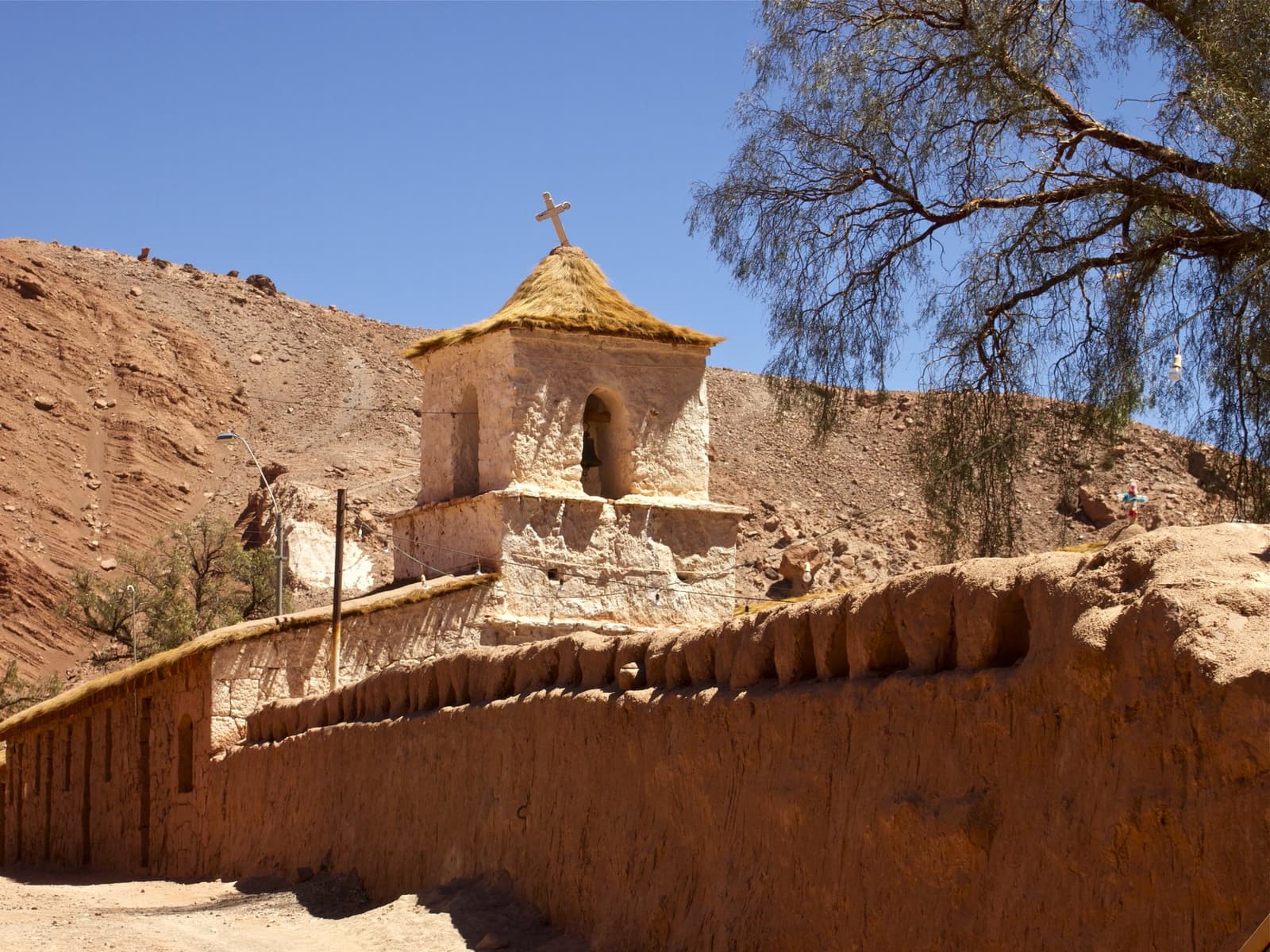 Oud genoeg voor een verhaal, klein genoeg om het te voelen. Photo: Igreja Rio Grande Een witgekalkte adobe klokkentoren met een rieten dak en stenen kruis verheft zich boven een leemstenen omheiningmuur langs de Atacama woestijn trekkroute, met rode rotshellingen en een diepe blauwe lucht op de achtergrond.