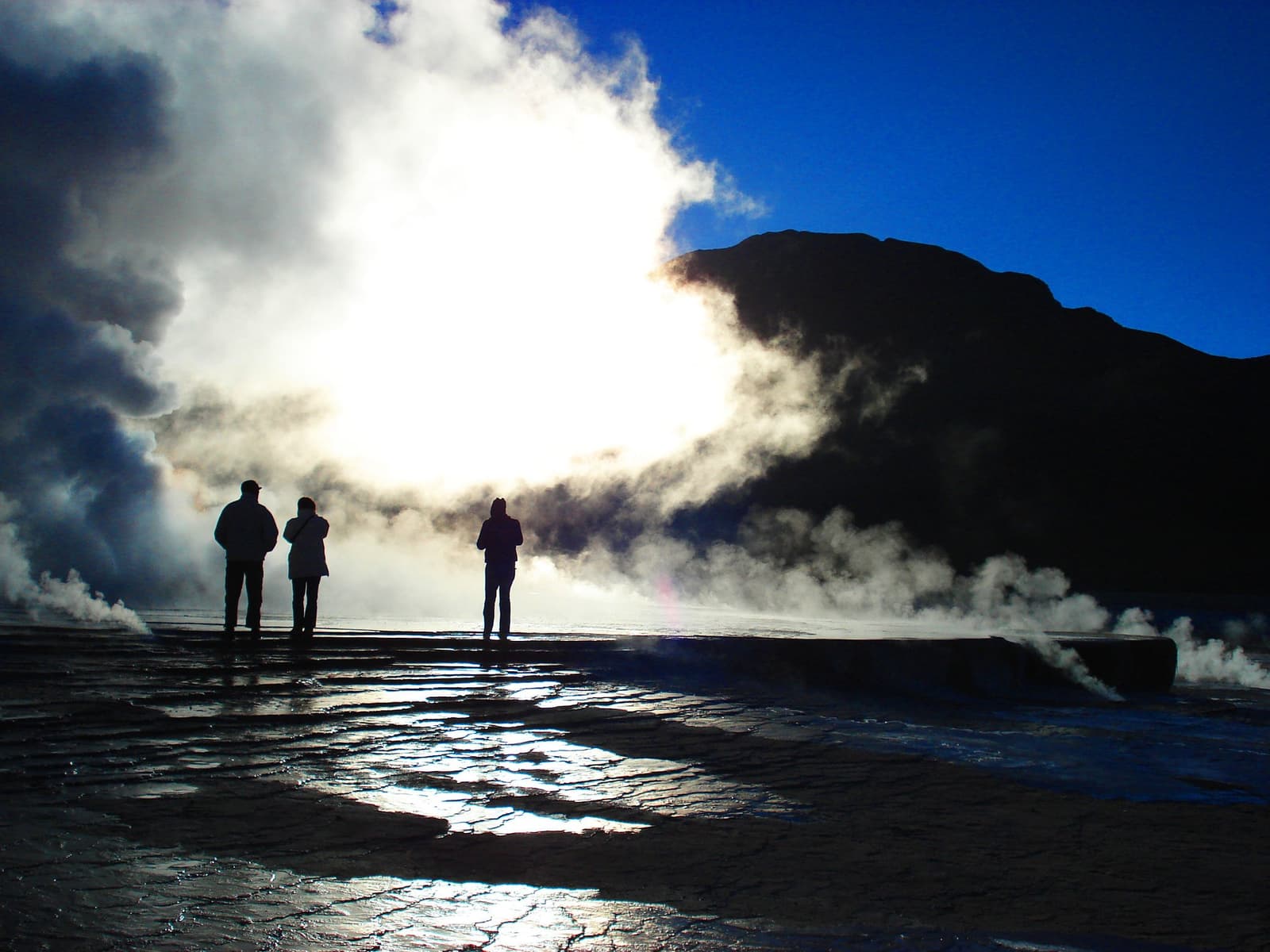 Stoom overal, ijskoude lucht in je gezicht, en de dag is nog maar net begonnen. Photo: Tatio Geysers Drie bezoekers als silhouet voor opstijgende geothermische stoomwolken van de El Tatio-geisers, met een donkere vulkanische bergkam en diepe blauwe lucht, tijdens een trektocht door de Atacamawoestijn