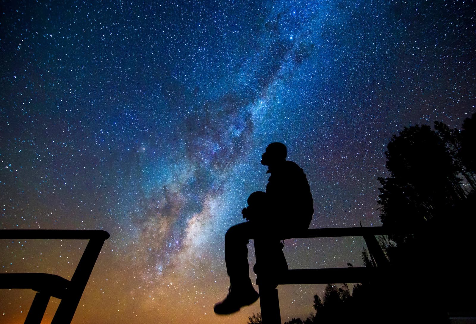 Dit soort nachten bewaar je ergens diep vanbinnen. Silhouet van een persoon zittend op een reling onder een heldere Melkweg tijdens een Atacama woestijn trek, met donkere boomcontouren aan de horizon